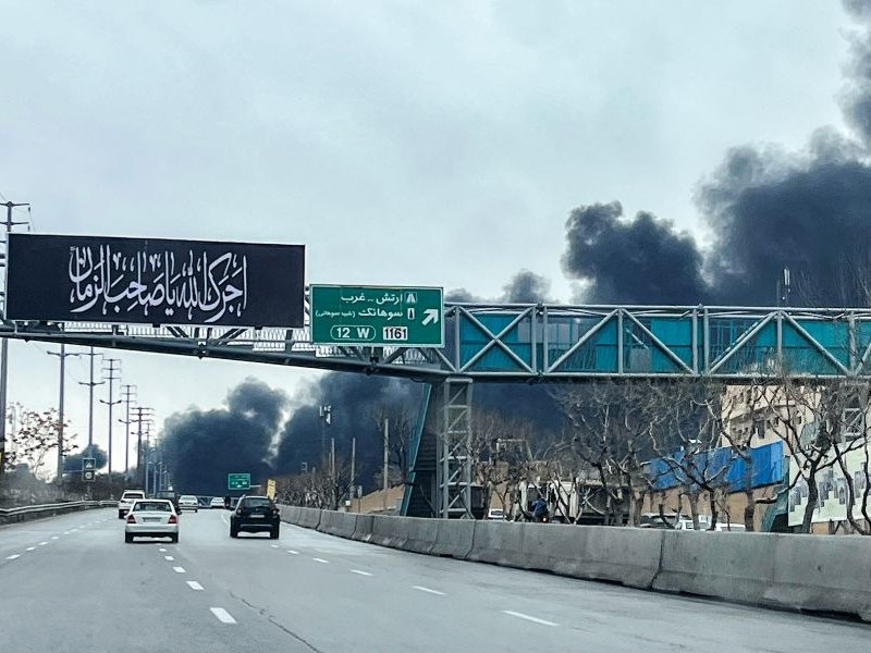 Vehicles move along a highway near plumes of black smoke billowing, in Tehran