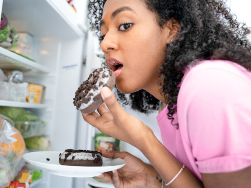A black woman about to bite into a donut standing at the fridge