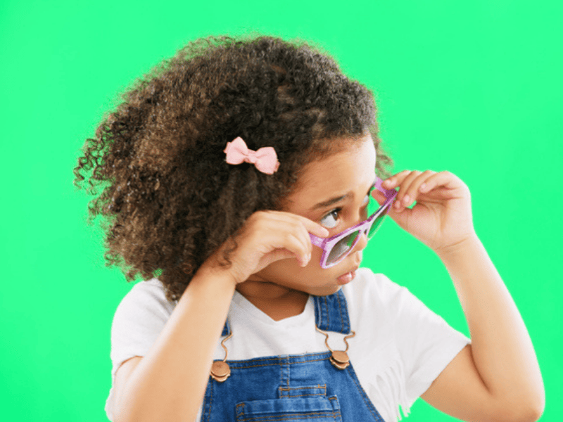 A child looking peaking through half pulled down sunglasses against a green screen