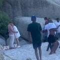 A couple embrace after their proposal on a beach and people dance around them