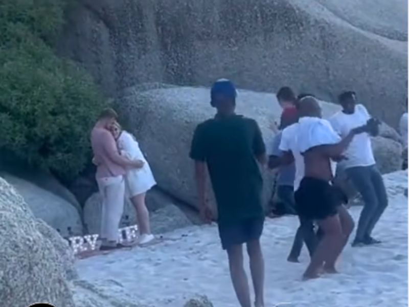 A couple embrace after their proposal on a beach and people dance around them