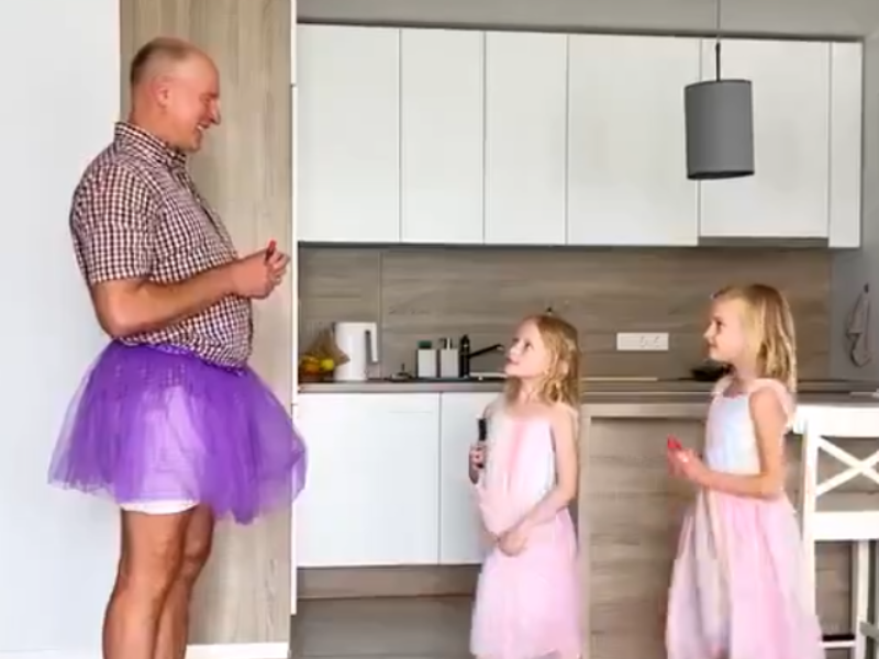 A father wearing a purple tutu stands with his two daughters