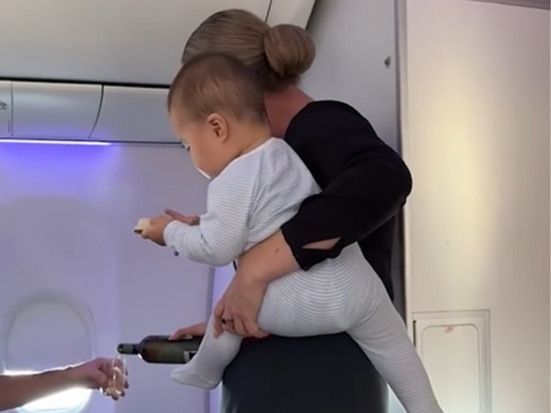 A flight attendant pours wine for a passenger while holding a baby