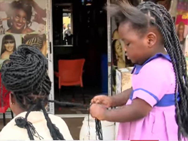 A little girl braids a womans hair outside a salon