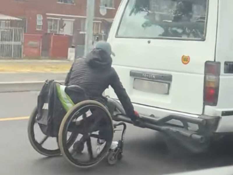 A man on a wheelchair holding onto the back of a moving taxi