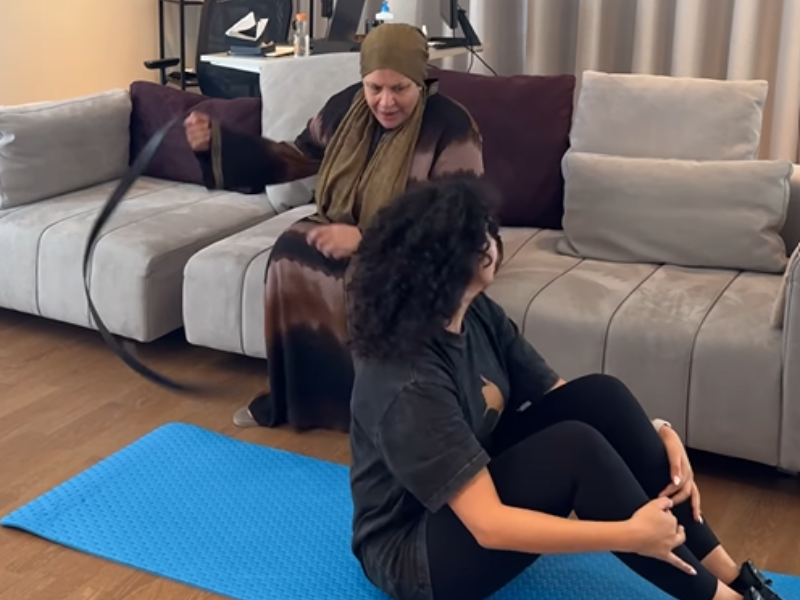 A mother whips a belt on the ground while her daughter does sit ups