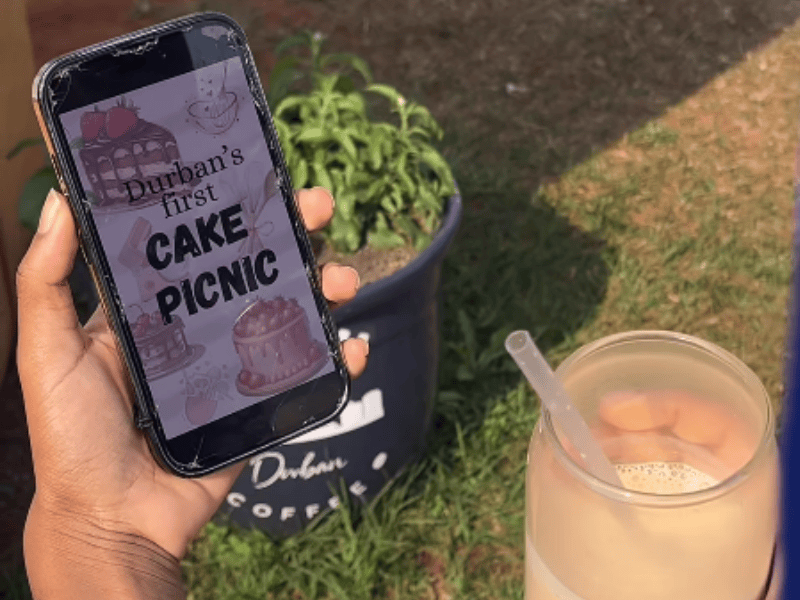 A person holds a phone showing a poster to Durban cake picnic and ice coffee