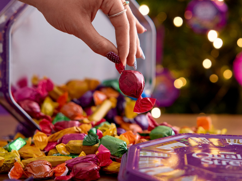 A person picks up a wrapped candy from tin of Quality Street