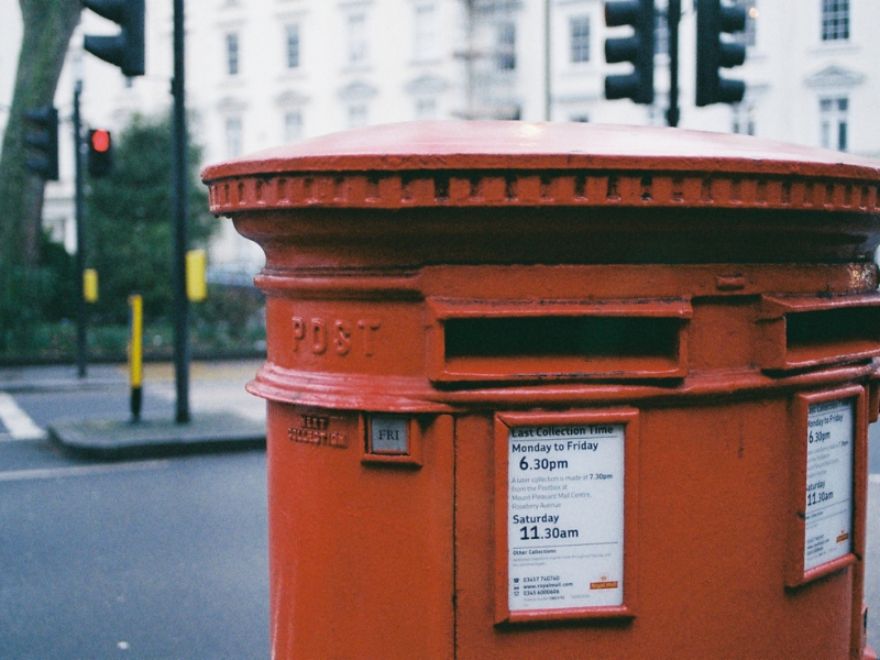 A reddish metal mailbox located on side of street