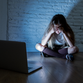 A sad girl holds her head in dismay while sitting opposite her laptop in a dark room