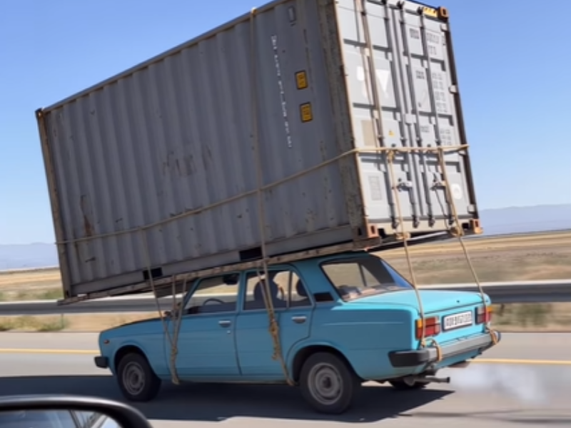A shipping container tied to the top of a car travelling on the road