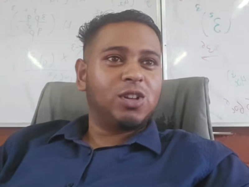 A teacher sitting at his desk with a whiteboard behind him