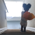 A toddler carries an orange sack over her shoulder and walks down stairs