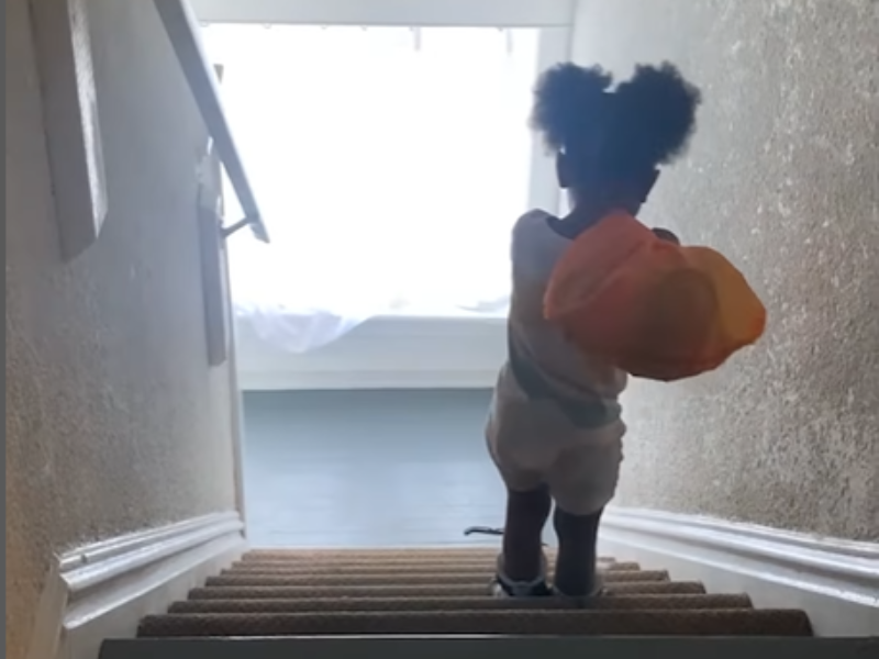 A toddler carries an orange sack over her shoulder and walks down stairs