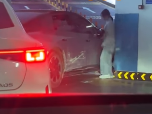 A woman tries to enter a car that is smashed on the side inside a parking lot