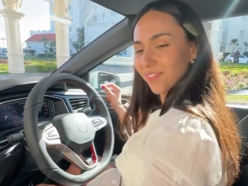 A woman wearing a white dress dances while sitting in the drivers seat