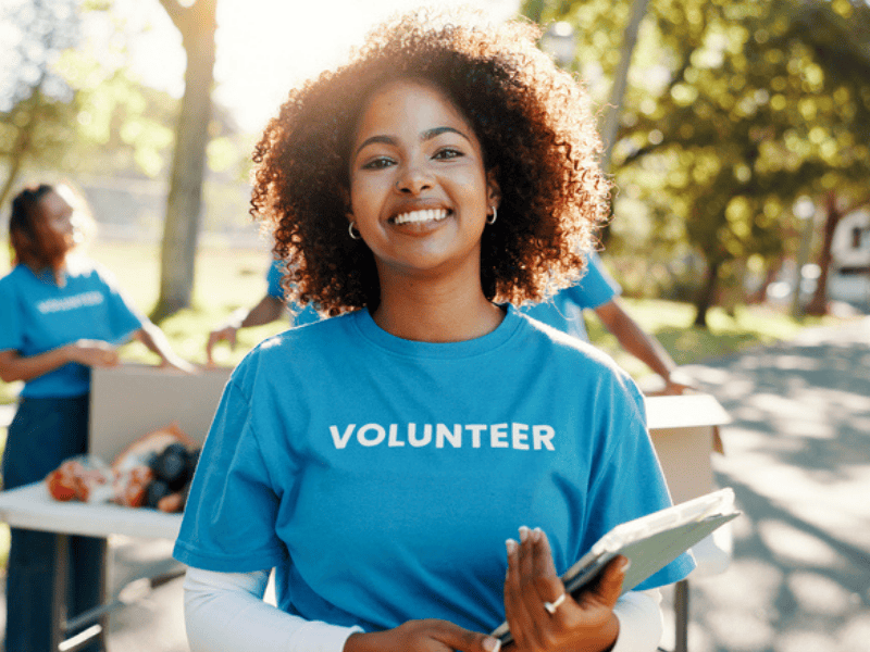 A woman with natural curly hair stands outdoors in front of a volunteer table