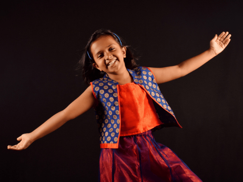A young girl dressed in Indian attire holds her arms out as if in a dance move