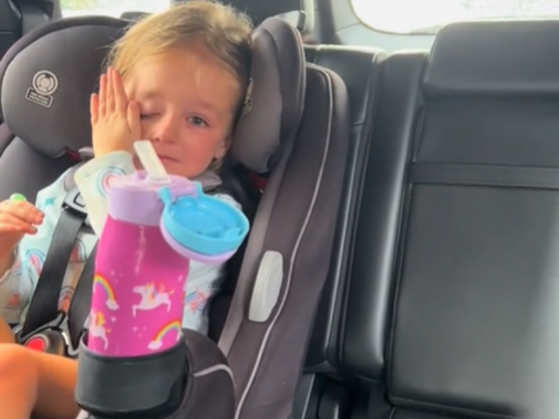A young girl is seated in her car seat wiping her tears away