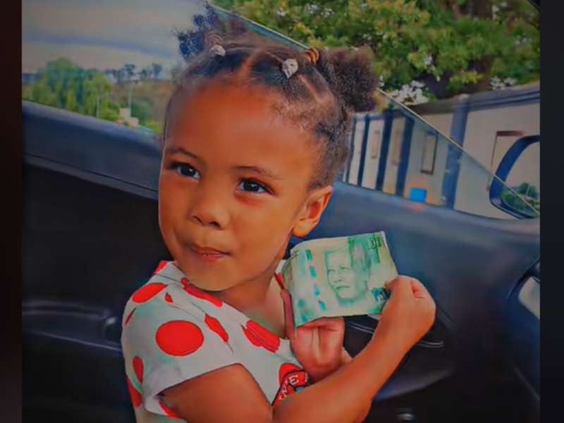 A young girl wearing a polka dot dress holds up a R10 ZAR note