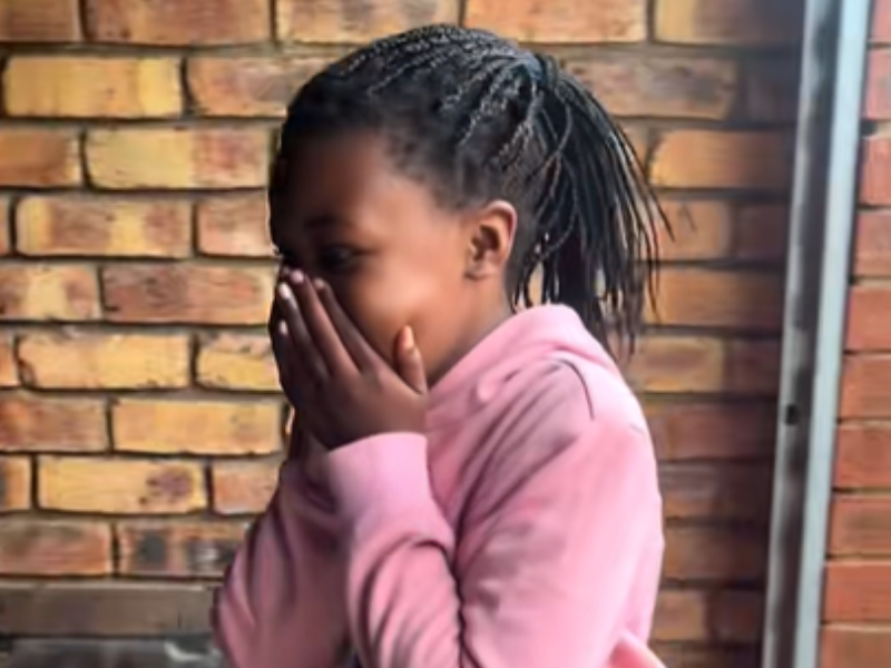 A young girl with braids covers her face in shock as she receives a surprise gift