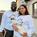 An interracial couple stand in front of their home with their baby
