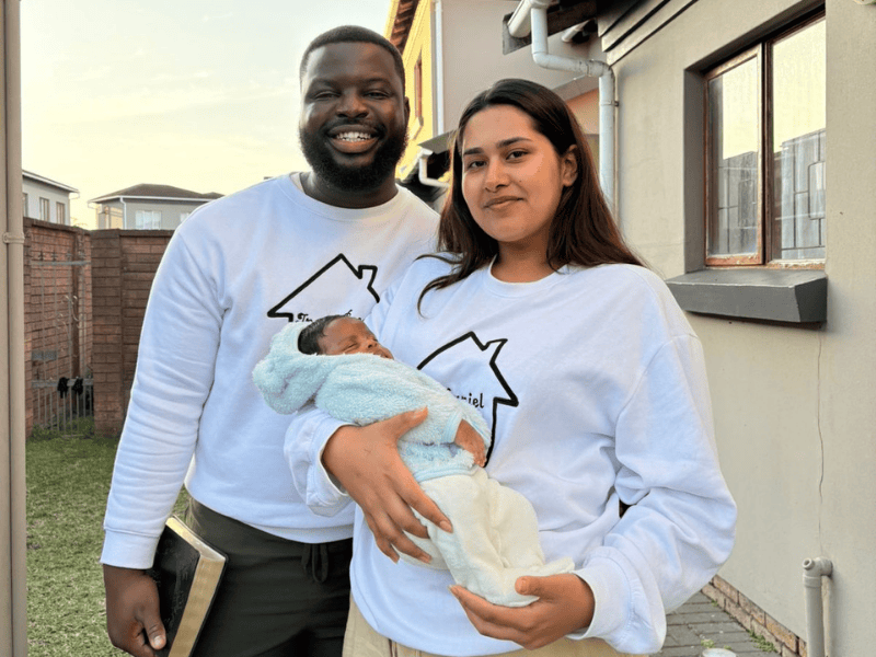 An interracial couple stand in front of their home with their baby