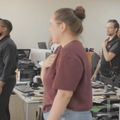 An office full of colleagues stand at their desks and sing the national anthem