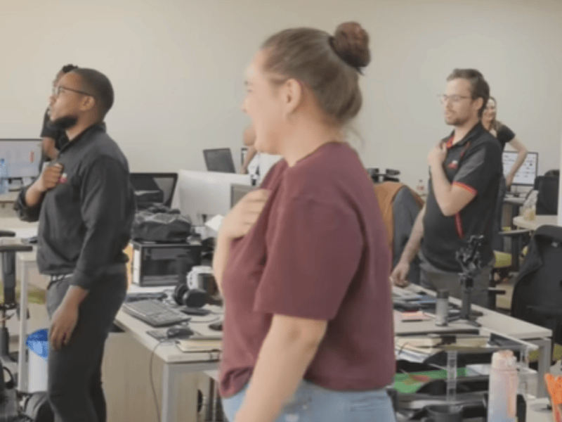 An office full of colleagues stand at their desks and sing the national anthem
