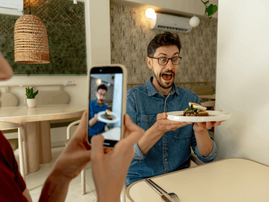 Excited man holds plate in restaurant while woman takes a photo.png