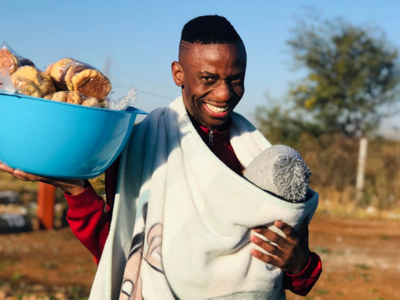 Father standing with a blue bowl of snacks and baby wrapped in blanket around his neck