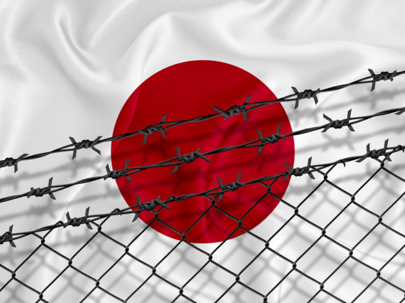 Japan flag behind a mesh fence and barbed wire indicating a prison