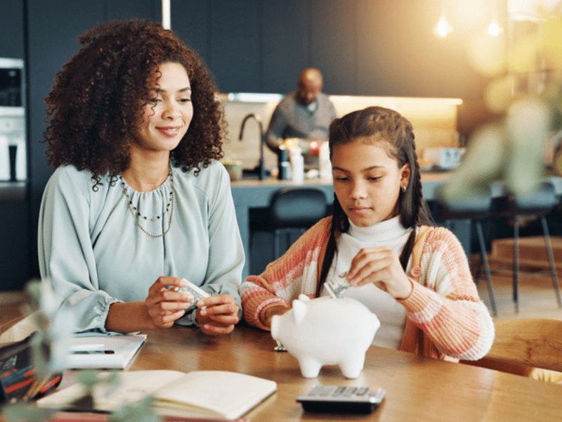 Mother and daughter sit at a kitchen table with a piggybank