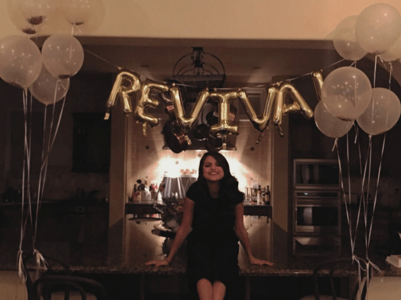 Selena Gomez sitting below gold letter balloons that read Revival
