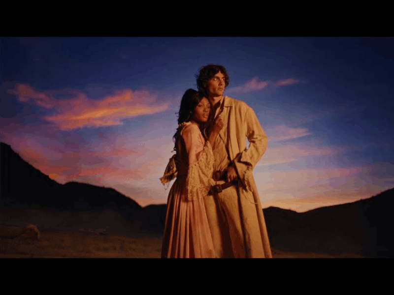 sombr embraces a woman while posing in an open landscape at dusk