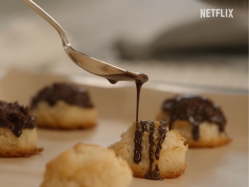 Spoon drizzles chocolate over coconut macaroons on a baking tray