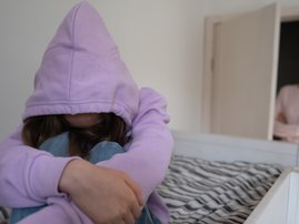 Teenager hiding in hood while sitting on a bed with adult standing in the background by the door