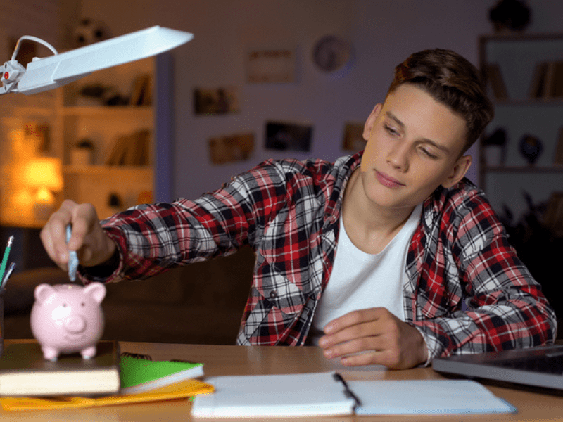 Teenage boy sitting at a desk places money into a pink piggyback
