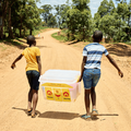 Two kids walk on a dirt road with a container filled with food supplies