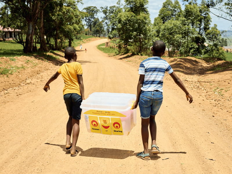 Two kids walk on a dirt road with a container filled with food supplies