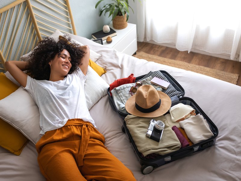 Woman lying down on bed next to open suitcase full of clothes, passport and hat