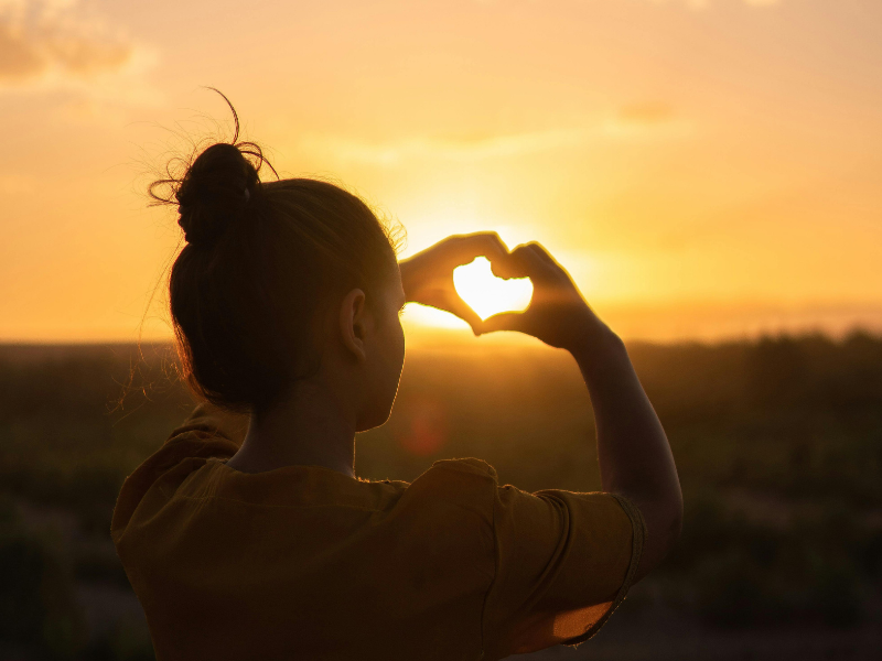 Woman sitting outdoors showing heart sign with hands during sunset