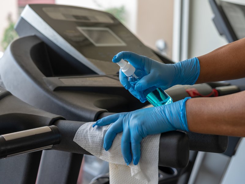 Staff using wet wipe and a blue sanitizer from the bottle to clean treadmill in gym