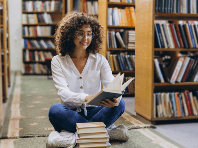 woman sits on the floor while reading books in a library
