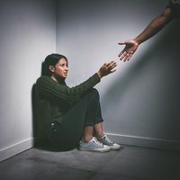 Young woman sitting in the corner of a dark room with a hand reaching out to help her